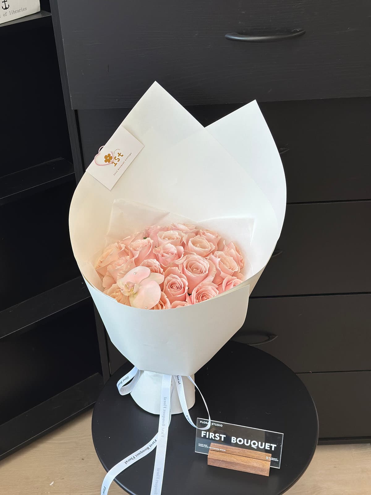 Blush pink roses in a tall white wrap, on a stand beside a First Bouquet sign.